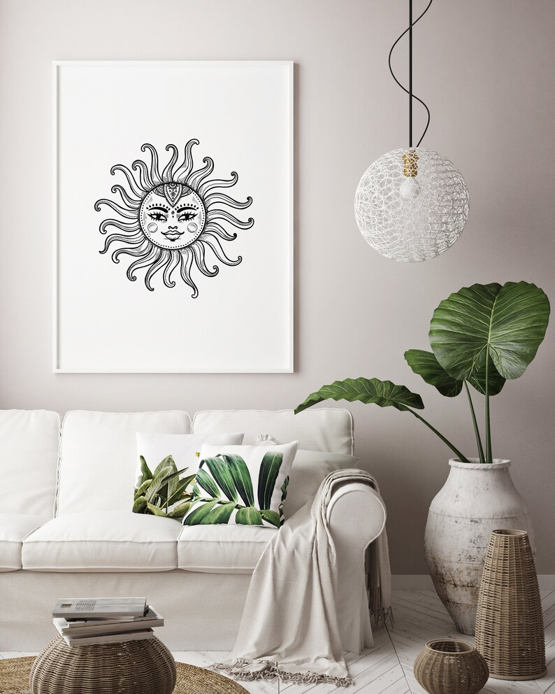 May include: A black and white illustration of a sun with a face, framed and hanging on a white wall above a white couch with green leaf patterned throw pillows.