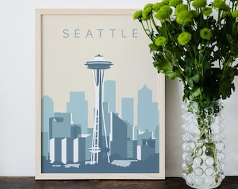 May include: A framed art print featuring a stylized Seattle skyline in shades of blue and white. The word "SEATTLE" is printed at the top. A vase of green flowers sits to the right of the print on a wooden surface.