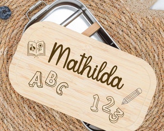 Peut inclure: Une boîte à lunch en bois avec un intérieur en acier inoxydable. Le couvercle porte une inscription personnalisée qui dit "Mathilda" avec des illustrations d'un livre, d'un crayon, de l'alphabet et de 123.