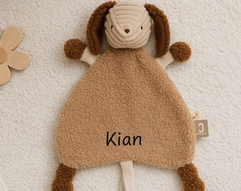 May include: A tan-coloured baby comforter in the shape of a dog, embroidered with the name "Kian". The dog features brown ears, paws, and a textured head. A wooden flower toy is visible in the background.