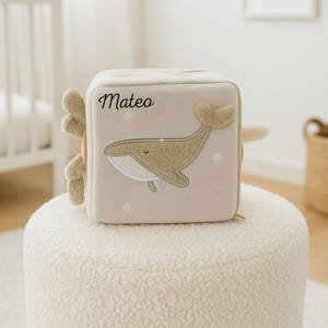 Può includere: Un cubo di stoffa beige con un'applicazione a balena e il nome "Mateo" in corsivo nero. La balena è marrone chiaro con un corpo strutturato e una pancia bianca. Il cubo ha bordi morbidi e arrotondati e piccoli dettagli circolari.