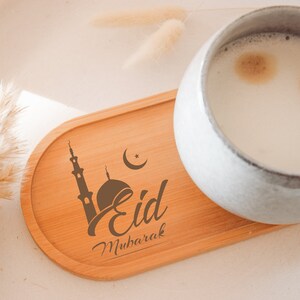 Könnte beinhalten: Ein Holz-Untersetzer mit der Aufschrift "Eid Mubarak" und einem Moscheedesign. Eine weiße Keramiktasse mit einem milchigen Getränk steht auf dem Untersetzer.
