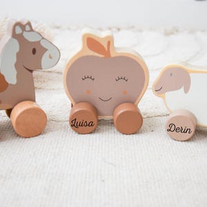 Könnte beinhalten: Holz-Nachziehtiere in Form eines Pferdes, Apfels und Schafs. Das Pferd ist braun und weiß, der Apfel ist beige mit einem Blatt und das Schaf ist weiß mit einem rosa Kopf. Jedes Spielzeug hat einen Namen auf seinen Rädern.