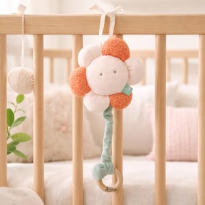 Può includere: Un mobile per bambini in peluche a forma di fiore, con petali bianchi e arancioni, appeso a una culla di legno. Il mobile ha uno stelo verde e un anello di legno con il nome "Anna". Un piccolo pompon è appeso alla culla.