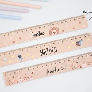 Könnte beinhalten: Drei Holzlineale, jeweils 50 cm lang, mit personalisierten Namen und dekorativen Elementen. Ein Lineal trägt den Namen "Sophie" mit einem Regenbogen, ein anderes "MATHEO" mit einer Rakete und das dritte "Amelie" mit floralen Akzenten.