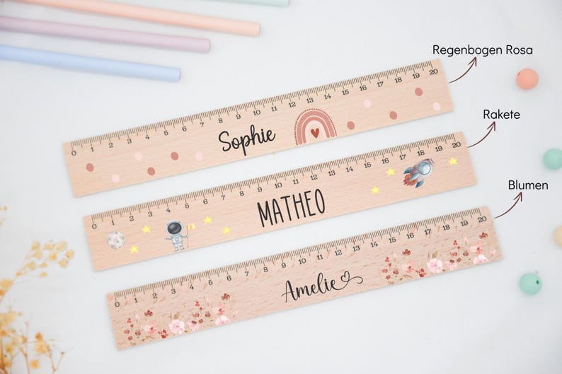 Könnte beinhalten: Drei Holzlineale, jeweils 50 cm lang, mit personalisierten Namen und dekorativen Elementen. Ein Lineal trägt den Namen "Sophie" mit einem Regenbogen, ein anderes "MATHEO" mit einer Rakete und das dritte "Amelie" mit floralen Akzenten.
