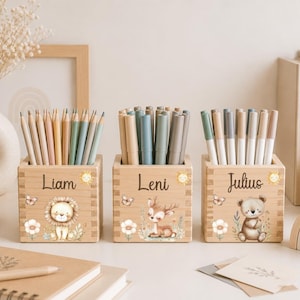 Peut inclure: Trois porte-crayons carrés en bois, chacun personnalisé avec un prénom : Liam, Leni et Julius. Chaque support présente une illustration imprimée d'un animal mignon—un lion, un cerf et un ours—ainsi que des fleurs, un papillon et un soleil.