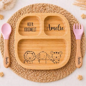 K&ouml;nnte beinhalten: Ein geteilter Holzteller mit einer Illustration von L&ouml;we, Elefant und Tiger, sowie einem rosa L&ouml;ffel und einer Gabel. Der Teller tr&auml;gt den Namen "Amelie" und den Schriftzug "Hier Kr&uuml;melt". L&ouml;ffel und Gabel tragen ebenfalls den Namen "Amelie".