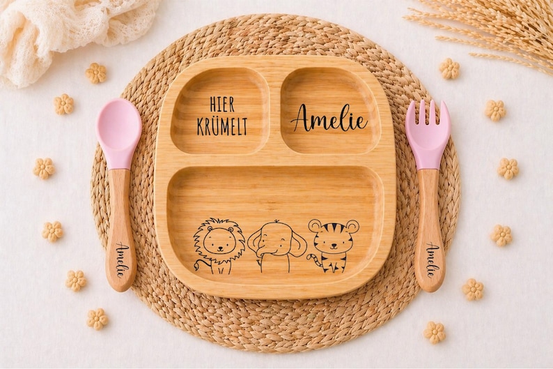 K&ouml;nnte beinhalten: Ein geteilter Holzteller mit einer Illustration von L&ouml;we, Elefant und Tiger, sowie einem rosa L&ouml;ffel und einer Gabel. Der Teller tr&auml;gt den Namen "Amelie" und den Schriftzug "Hier Kr&uuml;melt". L&ouml;ffel und Gabel tragen ebenfalls den Namen "Amelie".