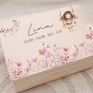 Op de afbeelding: Lichte houten bewaardoos versierd met roze en witte bloemmotieven. De doos is voorzien van de naam "Lina" en geboortegegevens. Een tekening van een meisje op een schommel staat erop. Een gehaakt paard is zichtbaar.