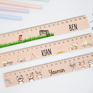 May include: Three wooden rulers, each 20 cm long, featuring custom names and illustrations. The top ruler shows a football scene with the name BEN. The middle ruler displays construction vehicles and the name KIAN. The bottom ruler features woodland animals and the name YASMIN.