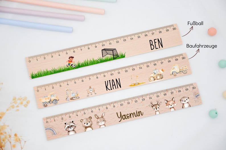 May include: Three wooden rulers, each 20 cm long, featuring custom names and illustrations. The top ruler shows a football scene with the name BEN. The middle ruler displays construction vehicles and the name KIAN. The bottom ruler features woodland animals and the name YASMIN.