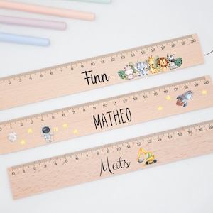 Könnte beinhalten: Drei Holzlineale, jeweils bis zu 20 Zoll (ca. 50 cm) messend, werden gezeigt. Die Lineale sind mit Namen und dekorativen Elementen wie einem Regenbogen, einer Rakete und einem Blumendesign personalisiert. Buntstifte in Pastellfarben und kleine dekorative Kugeln sind im Hintergrund.