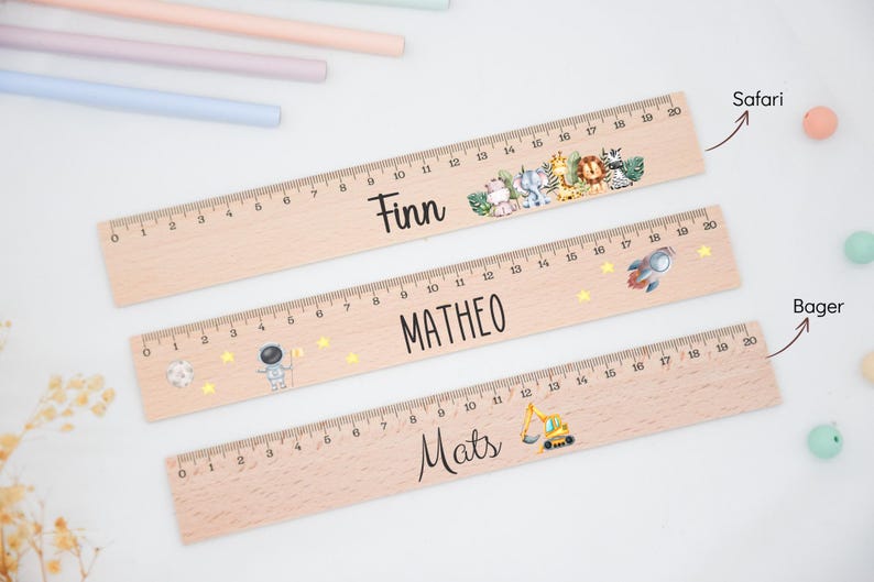 Könnte beinhalten: Drei Holzlineale, jeweils bis zu 20 Zoll (ca. 50 cm) messend, werden gezeigt. Die Lineale sind mit Namen und dekorativen Elementen wie einem Regenbogen, einer Rakete und einem Blumendesign personalisiert. Buntstifte in Pastellfarben und kleine dekorative Kugeln sind im Hintergrund.
