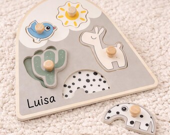 May include: Wooden puzzle with animal shapes, including a bird, sun, llama, cactus, and elephant. The puzzle is light gray with a wooden frame and knobs. The name "Luisa" is printed on the puzzle.