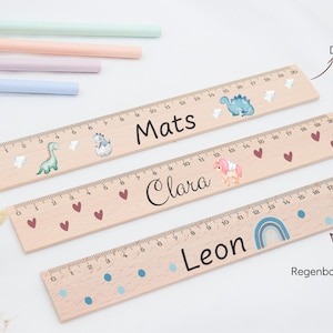 May include: Three wooden rulers with 20 cm markings. The top ruler features the name Mats with dinosaur graphics. The middle ruler displays the name Clara with heart graphics. The bottom ruler shows the name Leon with a blue rainbow graphic.
