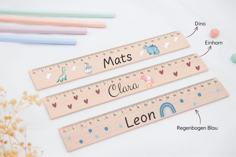 May include: Three wooden rulers with 20 cm markings. The top ruler features the name Mats with dinosaur graphics. The middle ruler displays the name Clara with heart graphics. The bottom ruler shows the name Leon with a blue rainbow graphic.