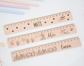 Puede incluir: Tres reglas de madera con nombres personalizados y diseños diferentes. La regla superior tiene un balón de fútbol y el nombre "Mats". La regla del medio tiene una sirena y el nombre "Mia". La regla inferior tiene flores y el nombre "Luisa".