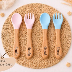 K&ouml;nnte beinhalten: Vier Baby-Essbestecke: zwei L&ouml;ffel und zwei Gabeln. Die L&ouml;ffel- und Gabelk&ouml;pfe sind rosa und blau, mit Holzgriffen. Die Griffe sind mit Namen graviert. Das Besteck ist auf einer gewebten Matte angeordnet.