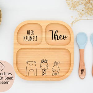 K&ouml;nnte beinhalten: Eine geteilte Holzplatte mit dem Namen "Theo" und dem Aufdruck "Hier Kr&uuml;melt". Die Platte zeigt Illustrationen eines B&auml;ren, einer Giraffe und eines Kaninchens. Enth&auml;lt einen blauen L&ouml;ffel und eine blaue Gabel mit Holzgriffen.