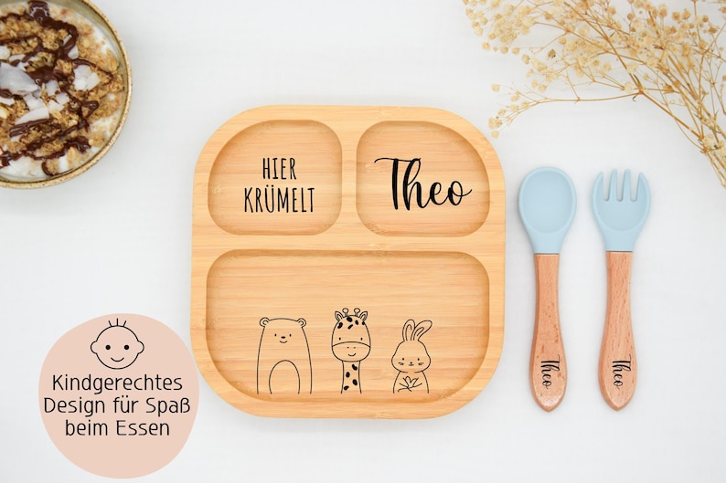 K&ouml;nnte beinhalten: Eine geteilte Holzplatte mit dem Namen "Theo" und dem Aufdruck "Hier Kr&uuml;melt". Die Platte zeigt Illustrationen eines B&auml;ren, einer Giraffe und eines Kaninchens. Enth&auml;lt einen blauen L&ouml;ffel und eine blaue Gabel mit Holzgriffen.