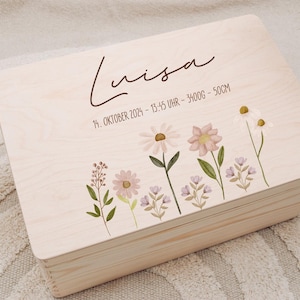 Könnte beinhalten: Helle Holz-Andenkenbox mit dem Namen "Luisa" in Schreibschrift. Die Box ist mit Aquarellblumen und Text verziert, der Geburtsdatum, Uhrzeit, Gewicht und Größe angibt. Ein gehäkeltes Pferdespielzeug ist sichtbar.