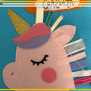 Puede incluir: Un unicornio de fieltro rosa con una crin morada y rosa, un cuerno de purpurina dorada y pestañas negras. El unicornio está sobre un fondo de fieltro azul con cintas de colores.