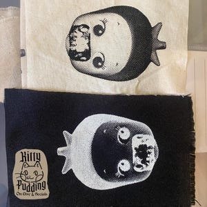 May include: Two fabric patches, one natural and one black, each featuring a white, stylized face with large eyes and a crescent-shaped head. The black patch has a Kitty Pudding logo with text: "On Etsy & Socials."