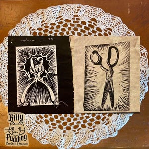 May include: Two linocut prints on fabric. One features a dental tool design, the other a pair of scissors. Both are black and white with a radiating burst design. The prints are on a white lace doily.