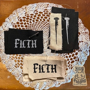 May include: A collection of fabric patches with the word "FILTH" in gothic lettering, and nail illustrations. The patches are black and beige, some with pins. The items are arranged on a white lace doily.