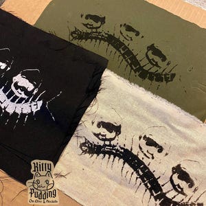 May include: Close-up of three fabric swatches with a repeating black and white graphic print of a creature's skeleton. The swatches are black, olive green, and off-white. The Kitty Pudding logo is visible.