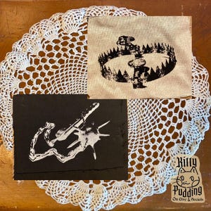 May include: Two patches with graphic designs. One patch is black with a white illustration of a weapon. The other patch is tan with a black illustration of a bear trap. The patches are on a white lace doily.