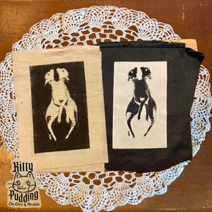 May include: Two fabric art pieces featuring a black and white dog illustration. One is on a beige background, the other on black. The dog illustration has a heart shape in the middle. The Kitty Pudding logo is in the bottom left corner.