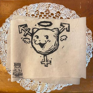 May include: A beige fabric square with a black linocut print of a cat with a halo, arrows, and a gender symbol. The text "Kitty Pudding On Etsy & Socials" is printed in black. The fabric is layered on a white lace doily.