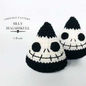 Può includere: Due ornamenti a forma di teschio di zucchero in crochet bianco e nero. Gli ornamenti hanno la forma di coni e hanno un sorriso cucito e occhi neri.