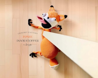 May include: Crochet pattern for a fox doorstop. The fox is orange and white with black eyes and a long, pink tongue sticking out. The fox is lying on its side, with its head and tail sticking out. The fox is holding a door open with its paws.