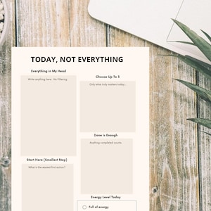 May include: A planner titled "TODAY, NOT EVERYTHING" with sections for writing, choosing tasks, and tracking energy levels. The planner is on a wooden surface with a laptop and plant.
