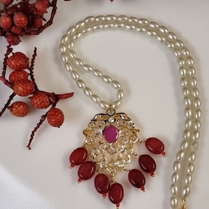 Pode incluir: Um colar com um pingente de ouro com uma pedra rosa cercada por pedras transparentes. O pingente está preso a um colar de pérolas brancas.