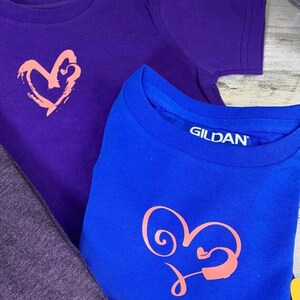 May include: Two t-shirts with a heart design. The top shirt is purple with a pink heart design. The bottom shirt is blue with a pink heart design and the text "GILDAN" on the tag.
