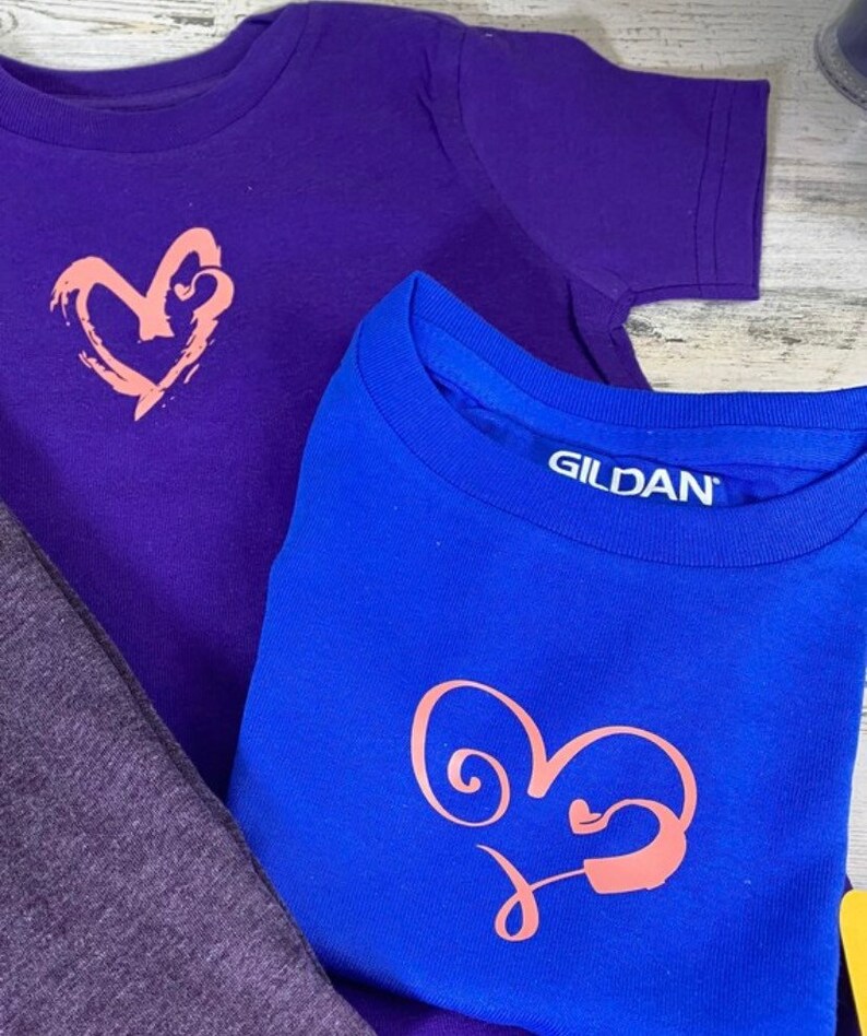 May include: Two t-shirts with a heart design. The top shirt is purple with a pink heart design. The bottom shirt is blue with a pink heart design and the text "GILDAN" on the tag.