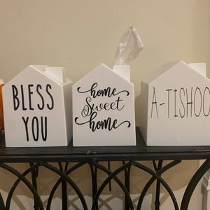 May include: Three white tissue box covers shaped like houses. The first says "Bless You", the second says "Home Sweet Home", and the third says "A-Tishoo".