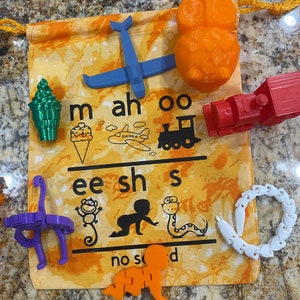May include: A yellow and orange fabric bag with a drawstring closure. The bag contains various 3D printed toys, including an airplane, an ice cream cone, an owl, a train, a monkey, a snake, a baby, and a chain. The bag also has a printed chart with the letters 'm', 'ah', 'oo', 'ee', 'sh', 's', 'no', 'so', and 'd'.