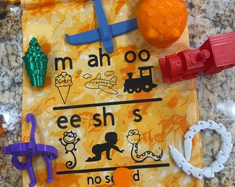 May include: A yellow and orange fabric bag with a drawstring closure. The bag contains various 3D printed toys, including an airplane, an ice cream cone, an owl, a train, a monkey, a snake, a baby, and a chain. The bag also has a printed chart with the letters 'm', 'ah', 'oo', 'ee', 'sh', 's', 'no', 'so', and 'd'.