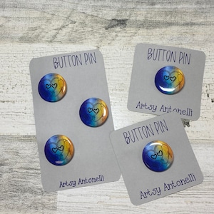 May include: Three button pins with a blue and yellow ombre heart design. The heart has a black outline and two small hearts inside. The pins are on white cards with the text "Button Pin" and "Artsy Antonelli" printed on them.