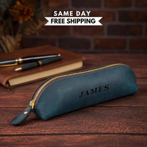 Pode incluir: Um estojo de couro azul com um zíper dourado e um puxador de couro. O nome JAMES está gravado em preto na lateral do estojo. O estojo tem um formato curvo e semicilíndrico e está posicionado sobre uma superfície de madeira escura.
