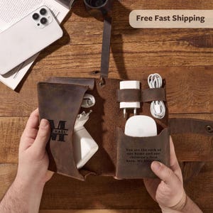May include: A brown leather tech organizer with compartments for chargers, cables, and a mouse. The organizer is personalized with the name "CHAEL" and a quote. A white smartphone is visible in the background.