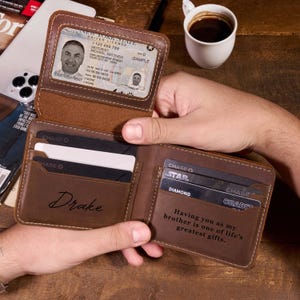 Puede incluir: Cartera de cuero marrón abierta con múltiples ranuras para tarjetas y una ventana de identificación transparente. La cartera está personalizada con el nombre "Drake" y la inscripción "Having you as my brother is one of life's greatest gifts."