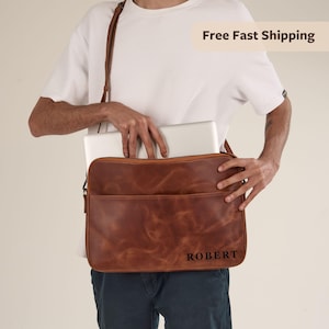 May include: A brown leather laptop bag with a shoulder strap. The bag has a front pocket and a zipper closure. The name "ROBERT" is printed on the lower right corner. A laptop is inside the bag.