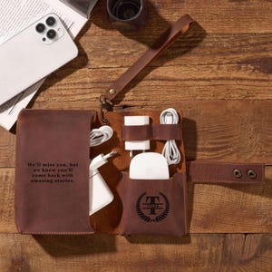 May include: Brown leather tech organizer with a wrist strap, open to reveal charging cables and adapters. The organizer has a pocket with the name "Tyler" and the text "We'll miss you, but we know you'll come back with amazing stories."