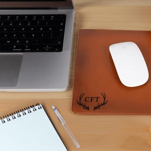 May include: A brown leather desk mat with a black deer antler design and the initials "CFT" in the center. A white wireless mouse is on the mat.